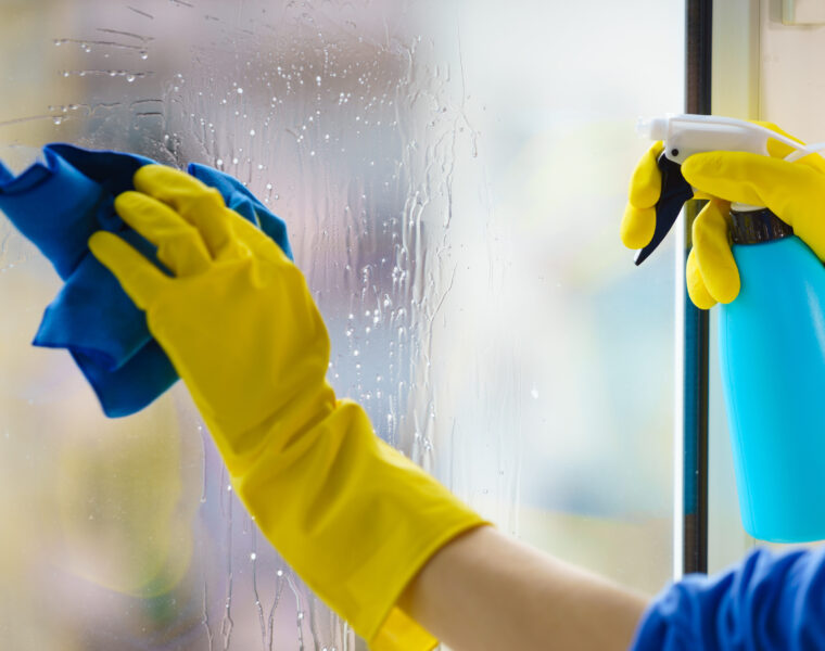 Bildausschnitt einer Aufnahme der Fensterreinigung mit gelben Handschuhen und blauem Putlumpen auf einem mit Reinigungsmittel besprayten Fenster.