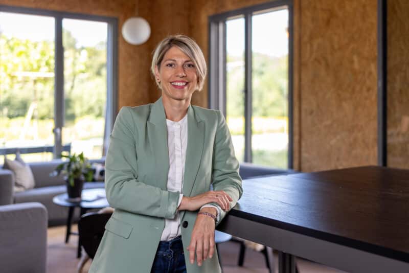 Frau mit blondem Kurzhaarschnitt im hellgrünen Blazer steht lächelnd an einem Tisch in einem modern eingerichteten Büroraum mit großen Fenstern.